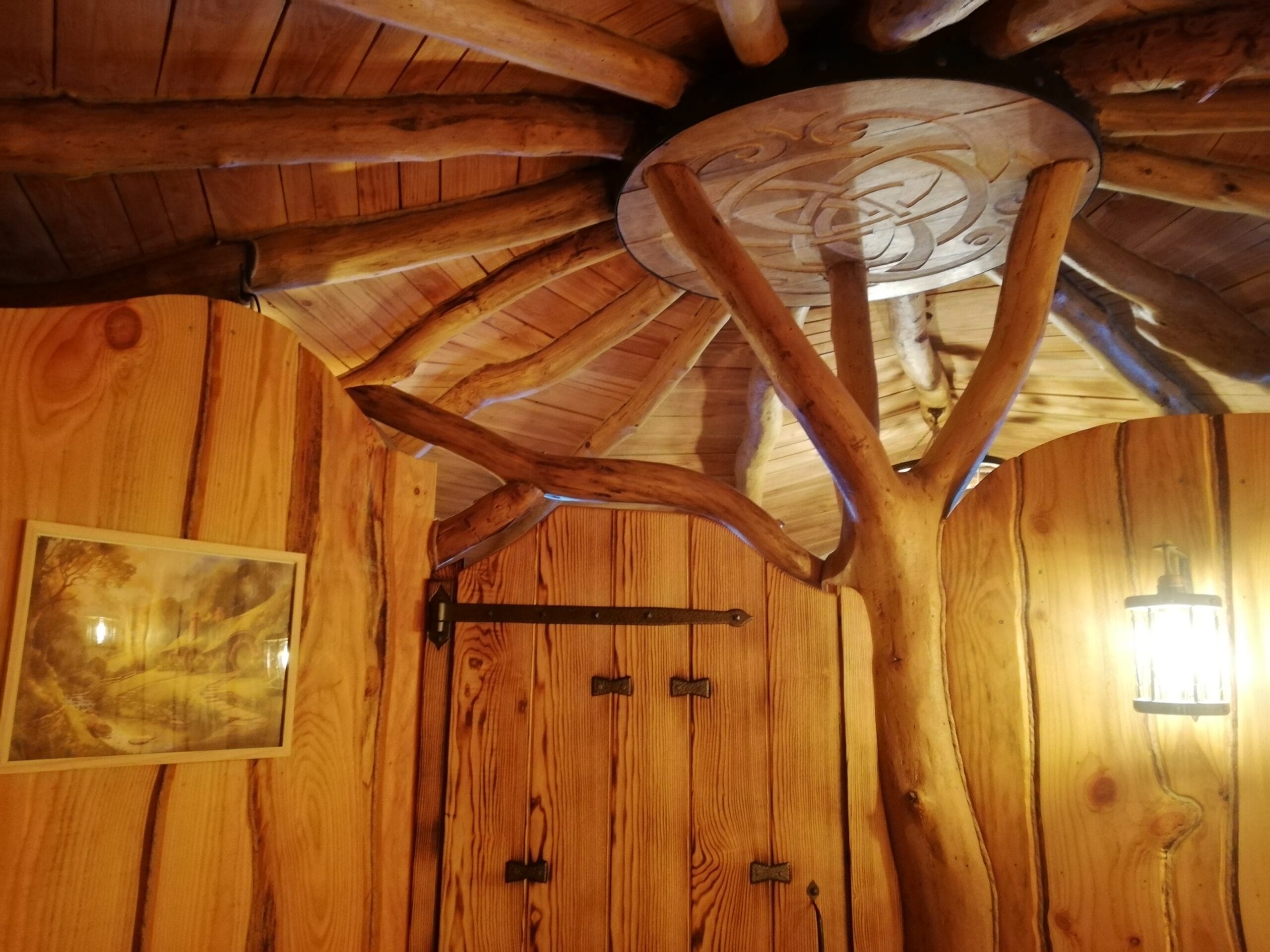 Hébergement Intérieur d’une cabane de Hobbit avec porte, luminaire et tronc de chêne central soutenant un disque en bois sculpté au plafond