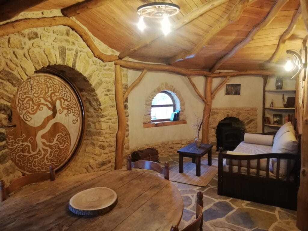 Salon d’une maison de Hobbit avec table, canapé fenêtre arrondie et porte ronde gravée à gauche