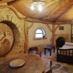 Hébergement Salon d’une maison de Hobbit avec table, canapé fenêtre arrondie et porte ronde gravée à gauche
